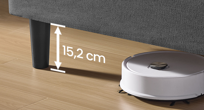 Aspirateur robotique circulaire blanc positionné sous un meuble gris. Le texte des mesures indique un espace de 15,2 cm entre le sol et le bas du meuble