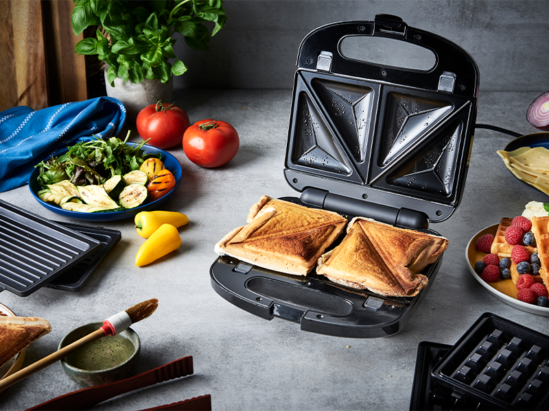 Séquence montrant une machine à sandwich noire en cours d'utilisation. Montre comment préparer et cuire un sandwich grillé avec des ingrédients et des garnitures visibles sur une surface de travail foncée.