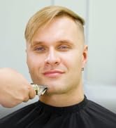 Gros plan d'une tondeuse à cheveux électrique utilisée sur les cheveux blonds d'une personne. La main du coiffeur est visible et tient la tondeuse