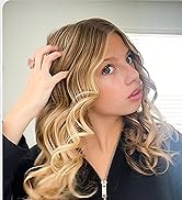 Femme blonde aux cheveux bouclés portant un haut zippé noir. Sa main est dans ses cheveux et met en valeur de volumineuses mèches blondes ondulées.