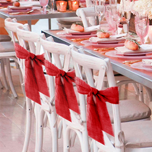Ensemble de table élégant avec des chaises blanches ornées de grands nœuds en ruban rouge. Les couverts comportent des assiettes de couleur corail et de la verrerie rose