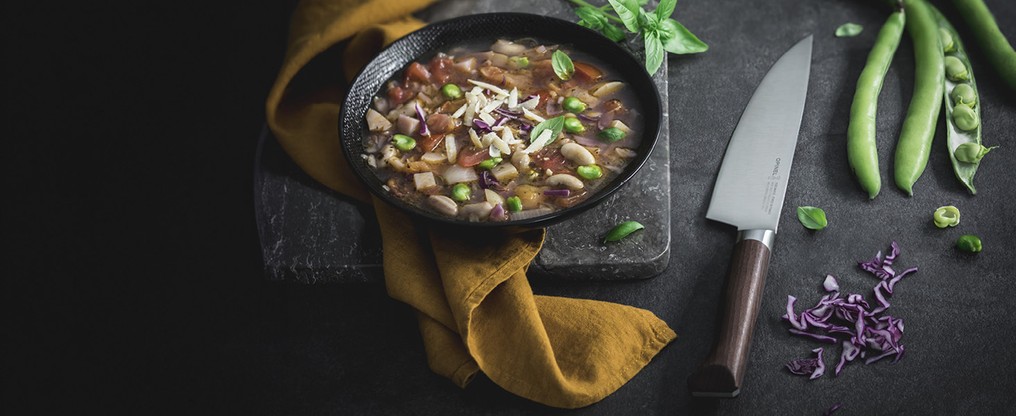 Photographie culinaire sombre et maussade d'une soupe ou d'un ragoût dans un bol en fonte, avec du chou violet, des pois verts et un couteau de chef sur une surface noire