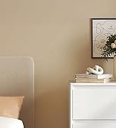 Coin d'une chambre aux murs beiges, à la commode blanche et à la décoration neutre. Art abstrait et arrangement végétal visibles. Vue partielle de la tête de lit et du coussin rembourrés