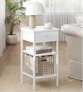 Table de chevet blanche avec tiroir, étagère inférieure et panier en osier. Lampe de table, tasse à café et livres visibles. Situé dans un salon lumineux.