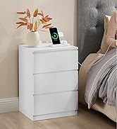 Table de chevet blanche à trois tiroirs, placée à côté d'un lit. Un vase avec des feuilles d'oranger et un smartphone en train de charger sur le dessus de la table.