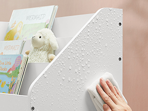 Gros plan d'une main touchant une surface texturée blanche avec des points en relief, probablement en braille. Peluche et livres visibles en arrière-plan.
