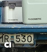 Deux glacières portables ou bacs de rangement de marque Dometic, l'un métallique et l'autre vert menthe, posés sur une surface bleue avec une plaque d'immatriculation visible
