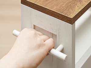 Gros plan d'une main ouvrant un tiroir d'armoire blanc avec un dessus en bois. Le tiroir est doté d'une simple poignée cylindrique blanche.