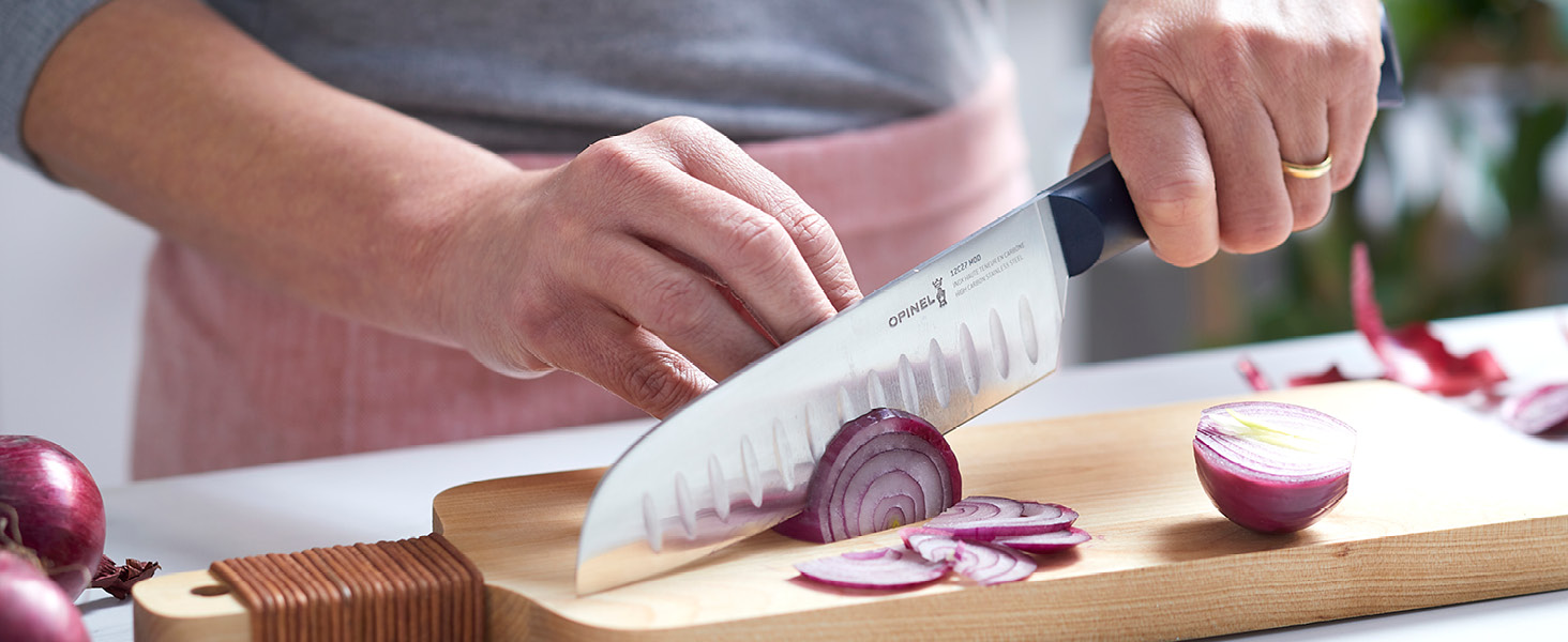 Les mains utilisent un couteau de chef pour trancher l'oignon rouge sur une planche à découper en bois, démontrant ainsi la technique de préparation des aliments en cuisine.