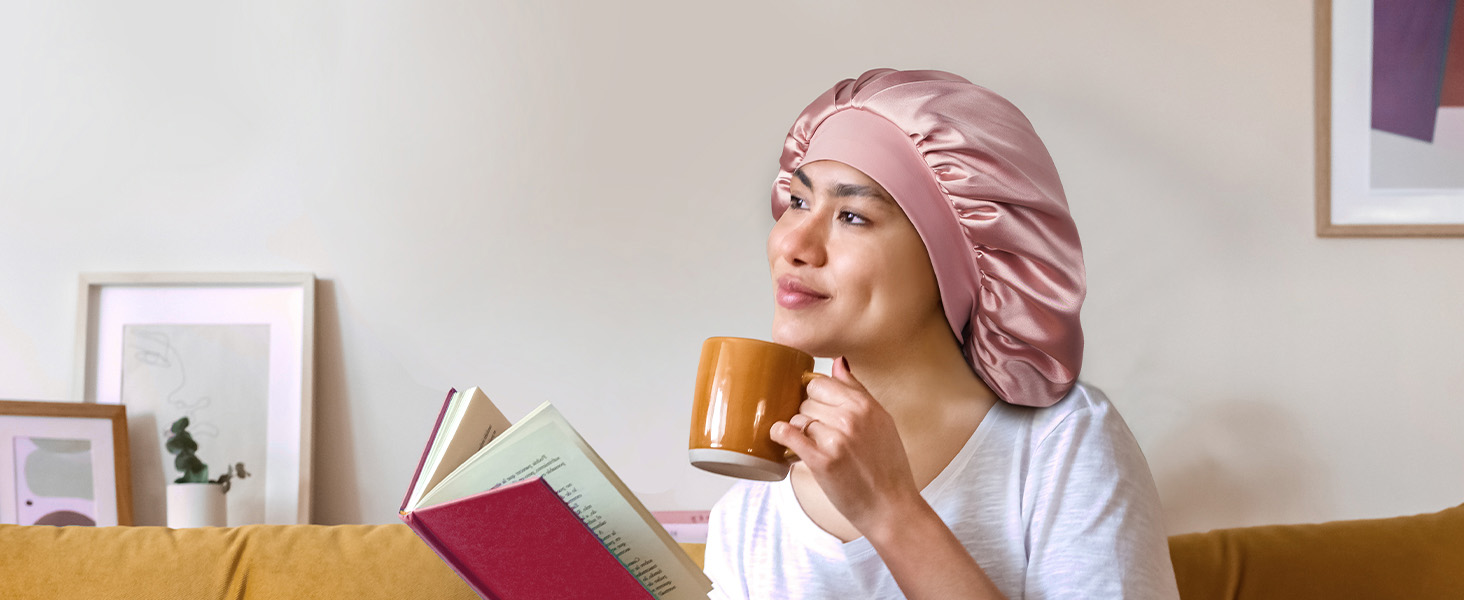 Bonnet de nuit en satin rose porté par une personne lisant un livre et tenant une tasse orange. Le bonnet est doté d'un bord élastique et d'un tissu froncé pour protéger les cheveux.