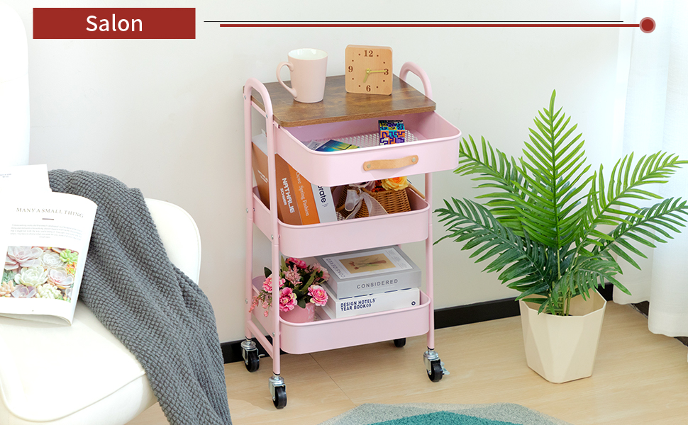 Chariot roulant rose à trois niveaux avec divers articles rangés à chaque niveau, positionné à côté d'une plante en pot dans une pièce lumineuse