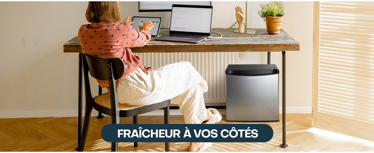 placé à côté d'un bureau avec une personne travaillant sur un ordinateur portable. L'unité est grise et noire, positionnée sur un plancher en bois près d'une fenêtre avec stores.