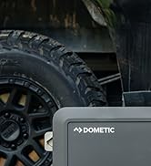 Gros plan d'un pneu tout-terrain robuste à bande de roulement profonde. Un appareil gris de la marque Dometic est partiellement visible au premier plan, avec une roue de véhicule en arrière-plan