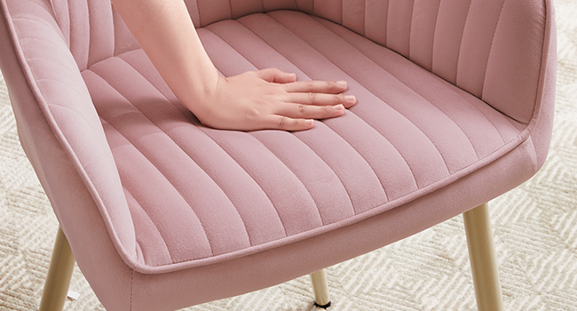 Gros plan d'une chaise en velours rose avec capitonnage à canaux verticaux, montrant une main posée sur un coussin de siège rembourré en peluche