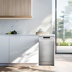 Intérieur de cuisine moderne avec armoires blanches, étagère supérieure en bois et lave-vaisselle mince en acier inoxydable. La grande fenêtre laisse entrer la lumière naturelle et illumine le comptoir avec des plantes