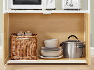 Étagère de cuisine ouverte en bois contenant un panier en osier, de la vaisselle blanche et des ustensiles de cuisine en acier inoxydable bien organisés.